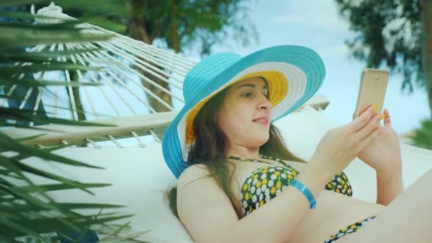 Una joven morena en bikini y con un sombrero de ala ancha brillante descansa en una hamaca y lee un libro en su teléfono móvil — Vídeos de Stock