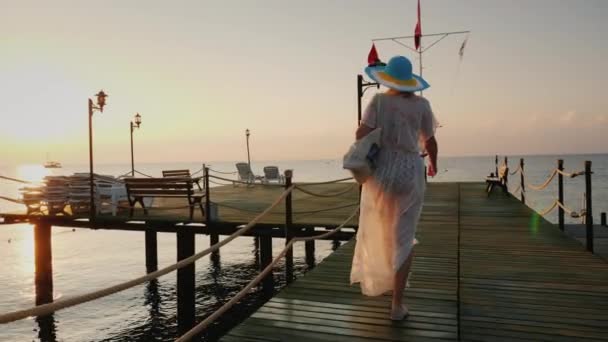 Bakifrån: En kvinna i par och hatt går på piren tidigt på morgonen. Andas in den friska luften. — Stockvideo
