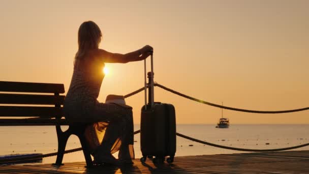 Een volwassen vrouw zit in een zomer sarafan op het strand, zit op een bank en houdt een koffer vast op wielen vroeg in de ochtend — Stockvideo