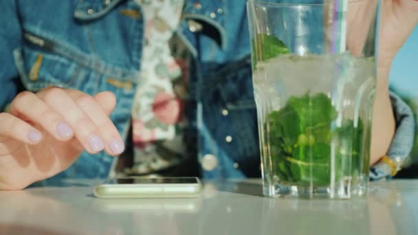 Les mains féminines feuilletaient les informations sur l'écran du smartphone. A proximité se trouve un verre avec un cocktail — Video