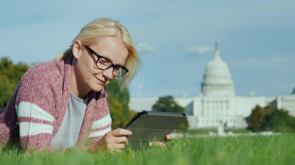 Gelukkige vrouw ligt op een grasveld tegen de achtergrond van het Capitool in Washington DC. Gebruikt een tablet. — Stockvideo