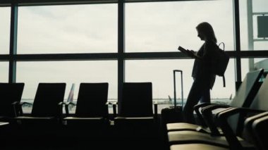 Geniş pencereli bir kadının silueti, tabletin tadını çıkarıyor. Terminal havaalanı, uçuşunuzu bekliyor.