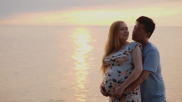 Щаслива молода багатонаціональна сім'я - азіатський чоловік обіймає свою вагітну дружину. На тлі заходу сонця над морем. — стокове відео