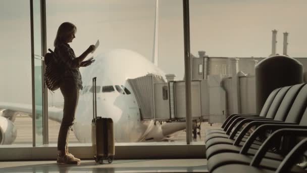 パスポートと搭乗券を持つ女性が空港の大きな窓に立っていて、窓の外には巨大な航空会社があります。旅の始まり — ストック動画