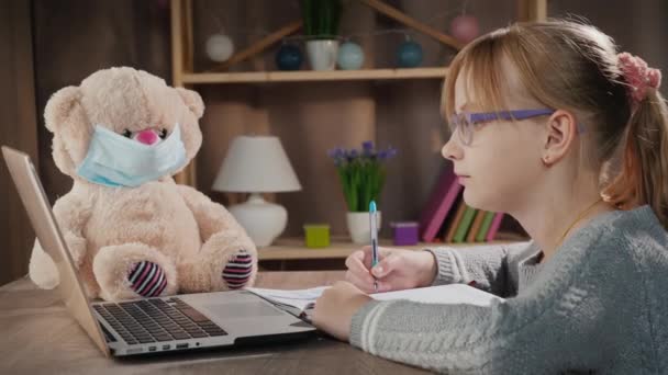 School online - leerling doet lessen in de buurt van laptop, naast een teddybeer met een beschermend masker — Stockvideo