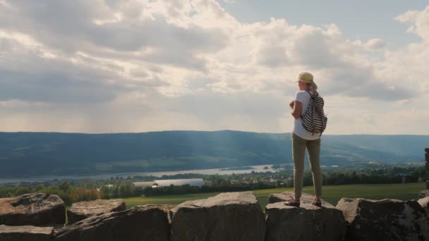 Žena s batohem stojí na staré kamenné zdi, dívá se do dálky na dramatickou oblohu a horizont — Stock video