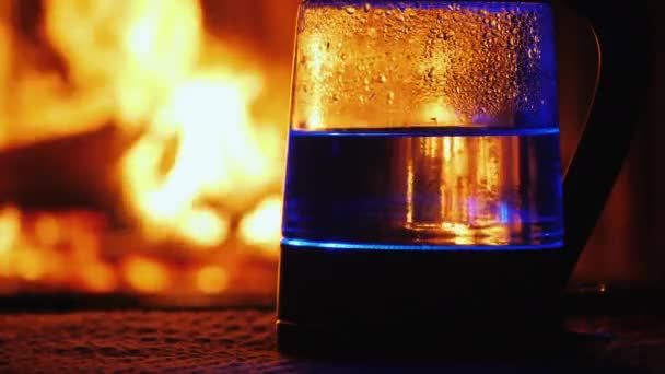 Το νερό βράζει σε ένα ηλεκτρικό βραστήρα, μια φωτιά στο παρασκήνιο καίει στο παρασκήνιο — Αρχείο Βίντεο
