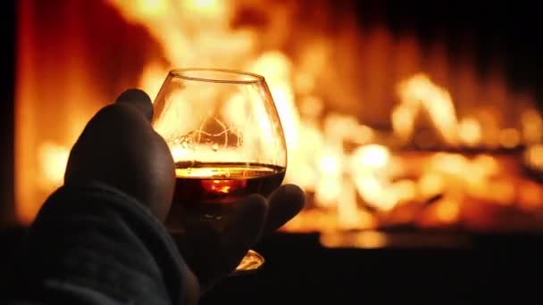 En man håller ett glas med alkohol i handen, elden från eldstaden reflekteras i drycken. Vinterkväll vid öppna spisen — Stockvideo