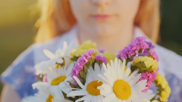 Chica rubia sostiene un ramo de flores, se encuentra en un prado al atardecer. Primer plano de tiro — Vídeo de stock