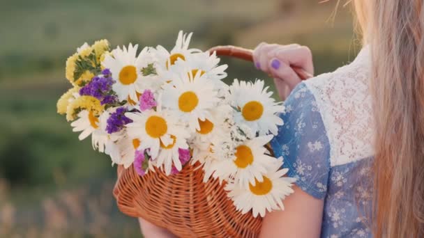 Vue arrière : Un enfant tient un panier de fleurs, se dresse dans un endroit pittoresque sur fond de prairies et de collines verdoyantes — Video