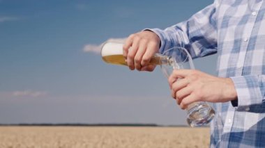 Bir adam şişeden bardağa bira dolduruyor. Tarladaki buğday kulaklarının ve mavi gökyüzünün arka planında