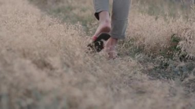 Parmak arası terlik giymiş orta yaşlı bir kadının bacakları yol boyunca otların arasında yürür.