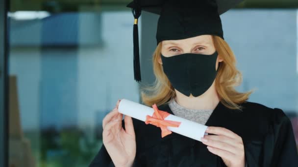 Een vrouwelijke afgestudeerde heeft een diploma. Ze draagt een badjas, een pet en een beschermend masker op haar gezicht.. — Stockvideo