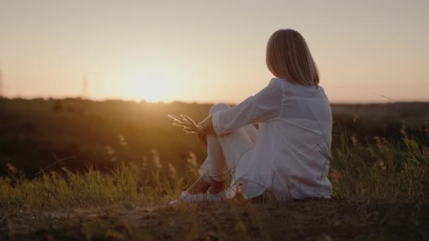 Женщина в белом костюме сидит на холме, любуясь закатом — стоковое видео