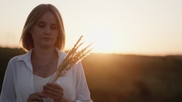 Portret kobiety trzymającej trawę polną stoi o zachodzie słońca w malowniczym miejscu — Wideo stockowe
