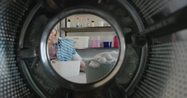 Уставшая женщина бросает одежду в стиральную машину. Сидя на полу в прачечной — стоковое видео