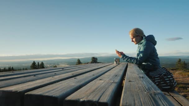 Eine Frau sitzt an einem Tisch, benutzt ihr Handy hoch oben in den Bergen Norwegens — Stockvideo