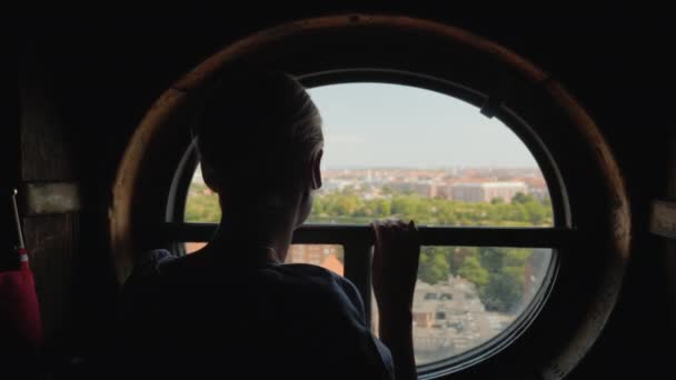 Vista trasera de una mujer mirando por una ventana redonda en los tejados de la ciudad de Copenhague en Dinamarca — Vídeo de stock