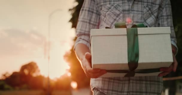 Een man draagt een mooi verpakt doosje met een geschenk, loopt door de straat in de zon — Stockvideo