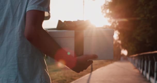 Achteraanzicht: Een man draagt een prachtig verpakt doosje met een geschenk, loopt door de straat in de zon — Stockvideo