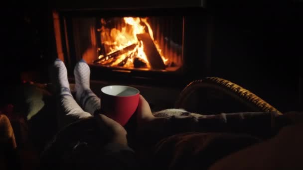 男は暖炉のそばで足を暖め、熱い飲み物で赤いカップを手に持っている — ストック動画