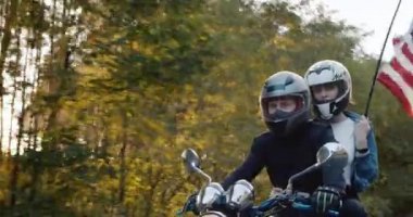 Kız arkadaşı olan genç bir motorcu güneşte motosiklet sürüyor, bir kadın ABD bayrağını tutuyor.