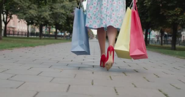 Widok z tyłu: Kobieta w czerwonych butach na wysokim obcasie niesie wielobarwne torby na zakupy ze sklepu z modą — Wideo stockowe