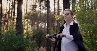 Okul üniformalı bir çocuk parkta scooter sürüyor, güneş ağaçların arasından parlıyor.