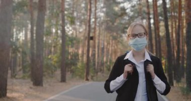 Maskeli okul çocuğu uzun ağaçlarla parkta yürüyor.