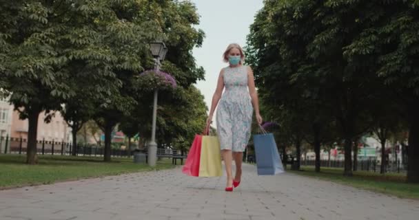 Una mujer con una máscara protectora camina por la calle, lleva bolsas de compras — Vídeo de stock
