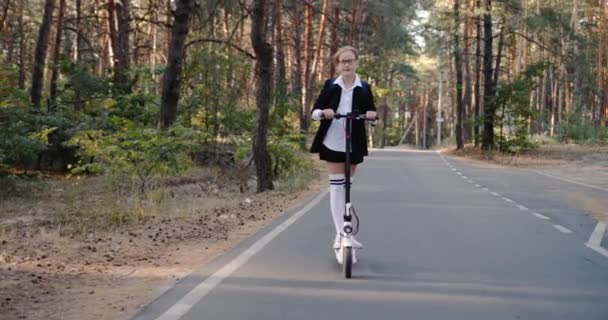 Un bambino in uniforme scolastica guida uno scooter attraverso il parco — Video Stock