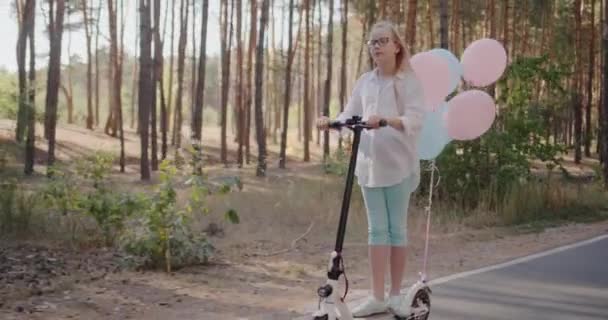 Un bambino su uno scooter decorato con palloncini cavalca attraverso il parco — Video Stock