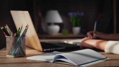 Bir kadın deftere yazar, dizüstü bilgisayarın yanındaki masaya oturur.
