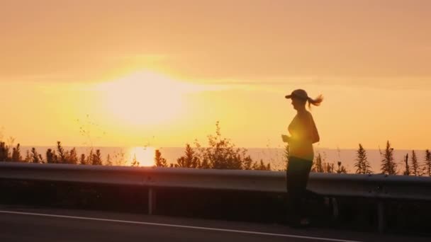 Атлетичная молодая женщина бежит вдоль побережья вдоль дороги. Спорт и здоровый образ жизни — стоковое видео