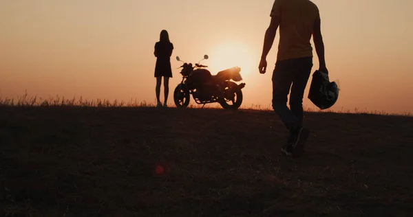 Die Silhouette eines Mannes, der zum Motorrad geht, wo er auf eine junge Frau wartet. Romantisches Reisekonzept — Stockfoto