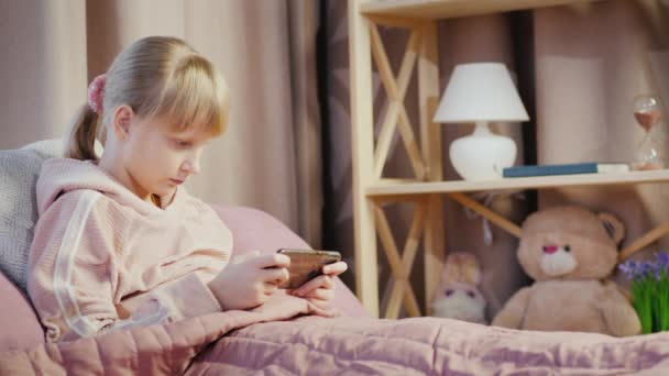 ブロンドの女の子は彼女の部屋でスマートフォンを使用します,ベッドに横たわっています — ストック動画