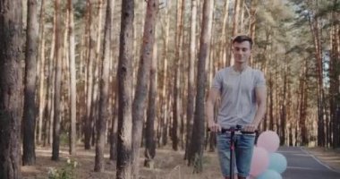 Genç bir adam parktan elektrikli scooter ile geçiyor, balonlar arkadan görünüyor.