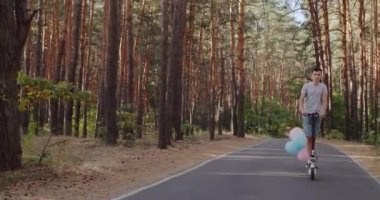 Genç bir adam parktan elektrikli scooter ile geçiyor, balonlar arkadan görünüyor.
