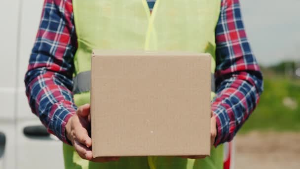 En kurir med en pappkartong står mot bakgrund av en vit leveransbil — Stockvideo