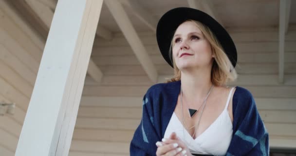 Retrato de uma mulher elegante em um chapéu e poncho, fica na varanda de sua casa de campo — Vídeo de Stock