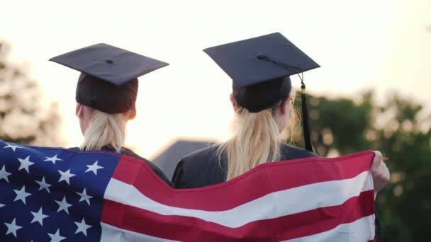 Две выпускницы колледжа с американским флагом на плечах, вид сзади — стоковое видео
