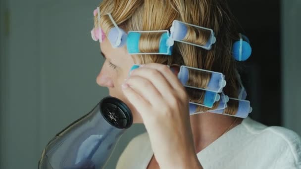 Mladá žena suší vlasy s fénem, kadeřnice na hlavě, aby dala tvar svému účesu — Stock video