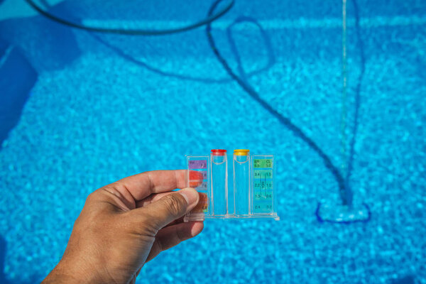 Рука держит набор для тестирования воды бассейна с уровнем pH и хлора над прозрачным голубым бассейном, показывая инструменты, используемые для обслуживания бассейна и контроля качества воды. Высокое качество фото