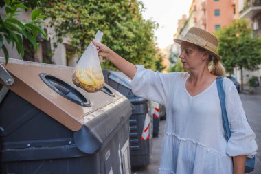 Hasır şapkalı ve beyaz elbiseli orta yaşlı bir kadın organik atıkları Roma 'da bir yerleşim bölgesinde geri dönüşüm konteynırına atıyor. Yüksek kalite fotoğraf