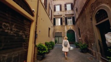 Beyaz elbiseli ve şapkalı bir kadın Roma 'da dar bir kaldırım taşı caddesinde yürüyor. Etrafı tarihi evler ve saksılarla çevrili.. 