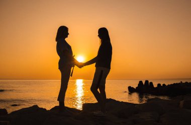 Gün doğumunda deniz kenarındaki kayalarda el ele tutuşan iki kadının silueti. Birbirlerine sıcak ve özenle bakıyor. Yüksek kalite fotoğraf