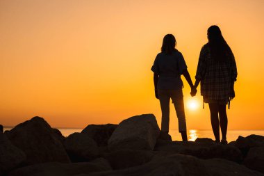Orta yaşlı bir kadın ve gün doğumunda deniz kenarında el ele tutuşan genç bir kadının silueti aile sevgisini, duygusal desteğini ve yeni bir günün ahengini simgeliyor. Yüksek kalite fotoğraf