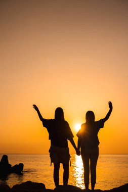 Gün doğumunda deniz kenarındaki kayalıklarda el ele tutuşan ve kollarını havaya kaldıran iki kadın yeni günü selamlıyor. Yüksek kalite fotoğraf