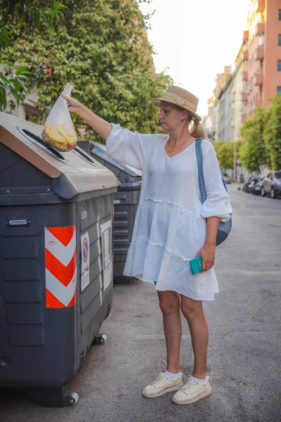 Hasır şapkalı ve beyaz elbiseli orta yaşlı bir kadın Roma 'da bir yerleşim bölgesinde belirlenmiş bir geri dönüşüm konteynırına organik gıda atıkları atıyor. Yüksek kalite fotoğraf