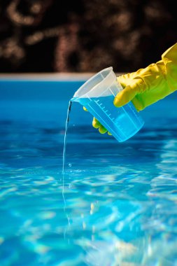 Sarı koruyucu eldiven, mavi kimyasal çözeltiyi ölçme kabından temizleme, dezenfeksiyon ve bakım için havuz suyuna döküyor. Yüksek kalite fotoğraf